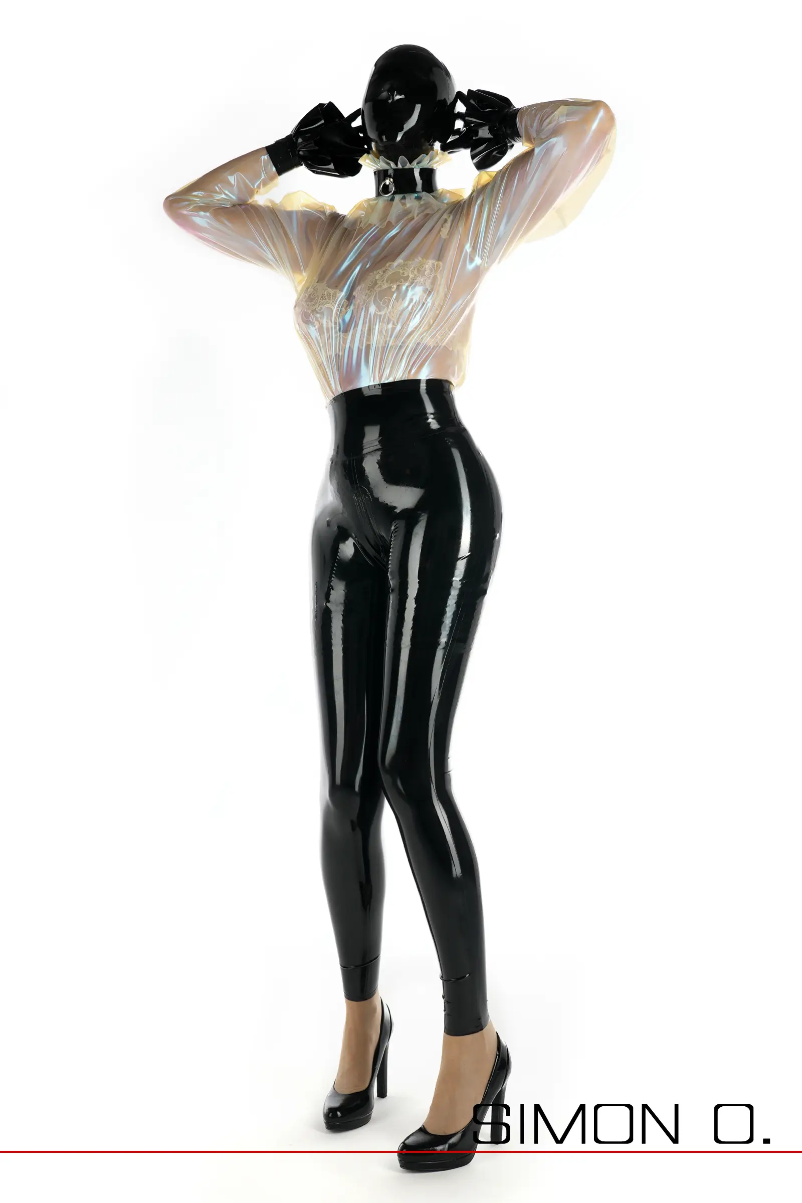 Eine Frau in einem Latex-Ganzanzug mit einem schillernden, durchsichtigen Oberteil im Blusenstil, High Heels, Handschuhen und einer Maske steht vor einem weißen Hintergrund.
