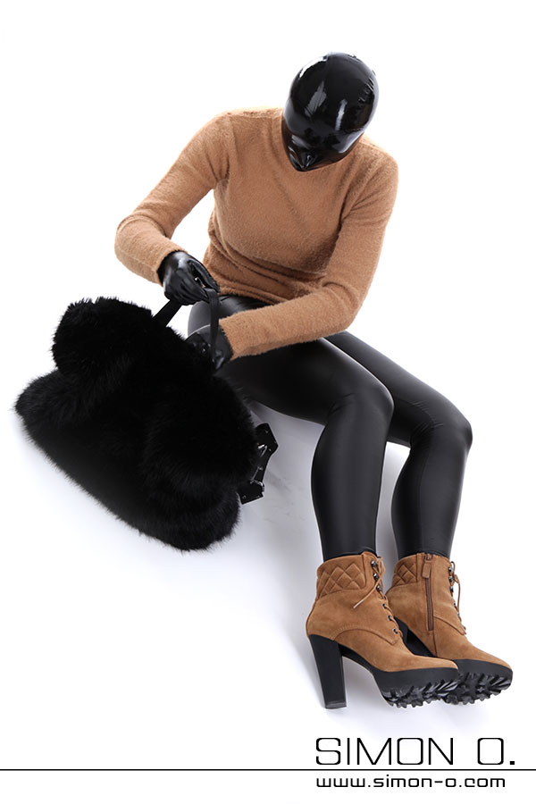Sexy Latex outfit unter Strassenkleidung. Eine Person in einem braunen Pullover, Latexkleidung und hellbraunen Stiefeln mit hohen Absätzen sitzt auf dem Boden und hält eine große, schwarze, flauschige Tasche.