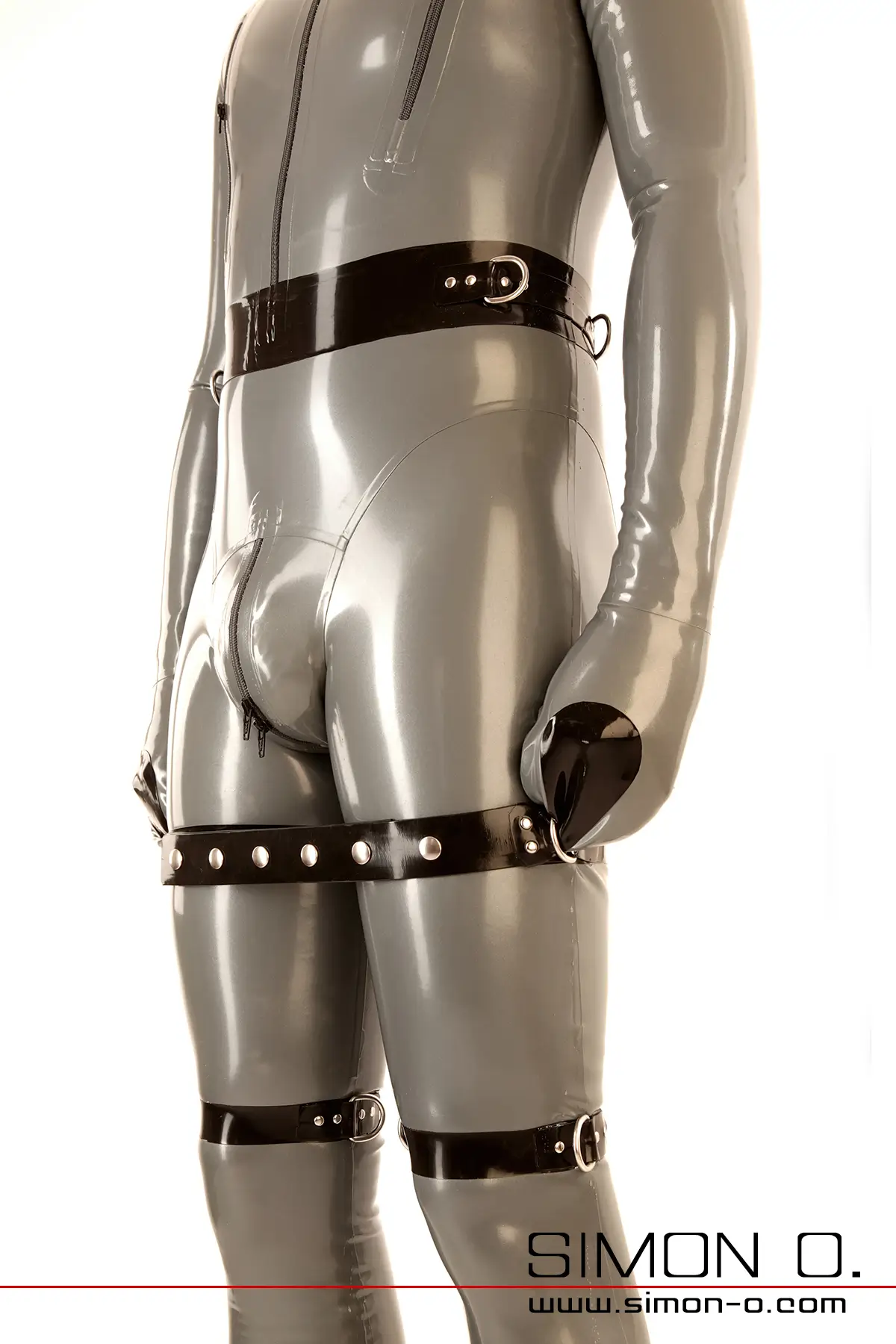 A man in a grey bondage latex suit with mittens has his arms fixed to his thighs and stands in front of a white background.