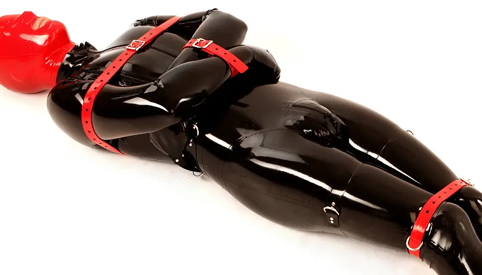 Black latex bondage catsuit A person in a black latex bondage catsuit and a red mask is lying on his back, arms and legs are bound with red straps on a white surface.