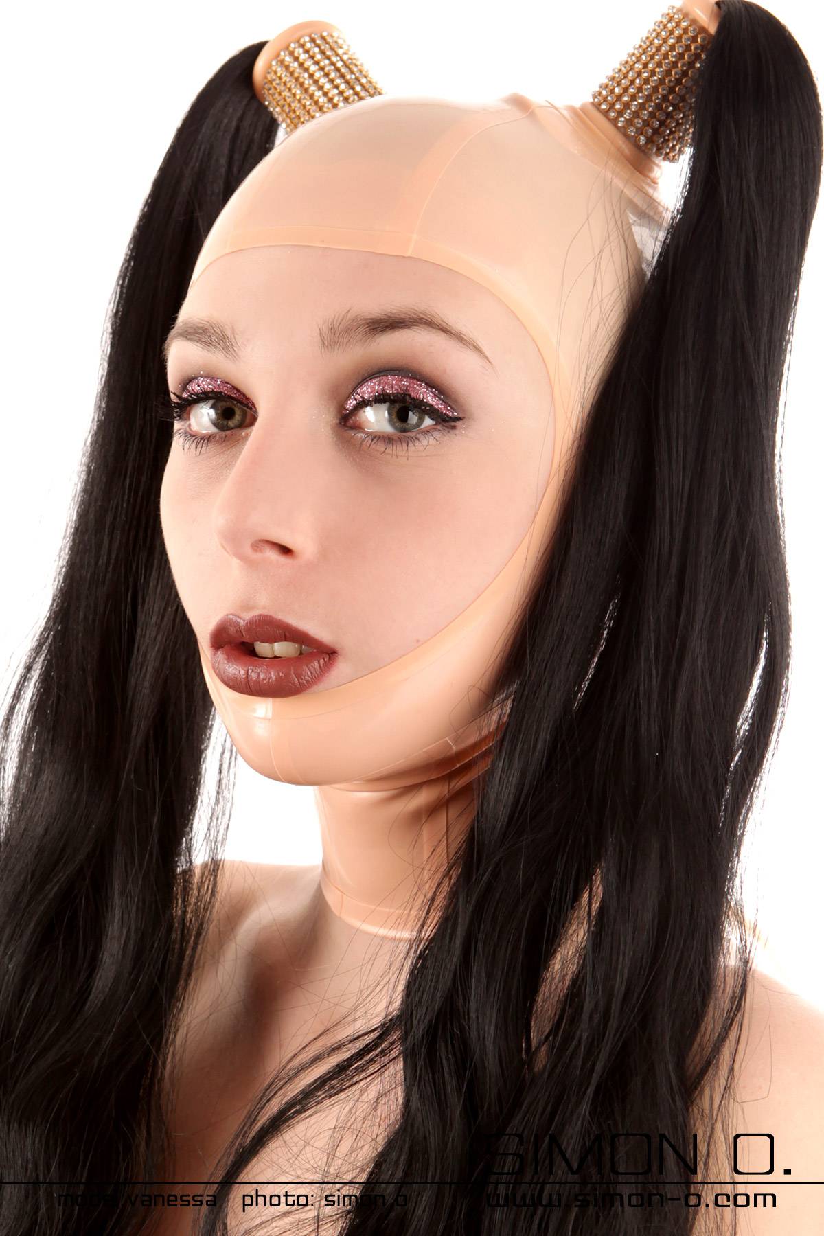 Geischtsoffene Latex Maske mit 2 Haarteilen Eine Person trägt eine hautfarbene Latex Maske mit offenen Gesichtsfeld und zwei daran befestigten schwarzen Haarteilen.