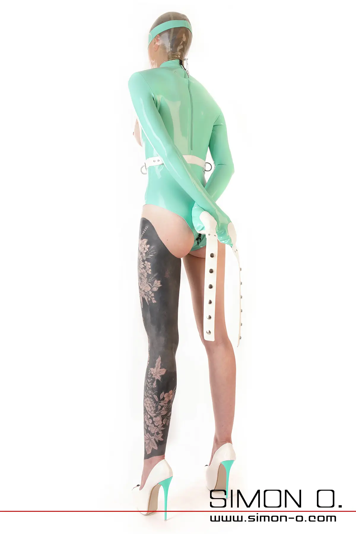 Eine Person in einem Latex-Bondage-Body, einer durchsichtigen Maske und High Heels steht mit gefesselten Armen vor weißem Hintergrund von hinten gesehen.