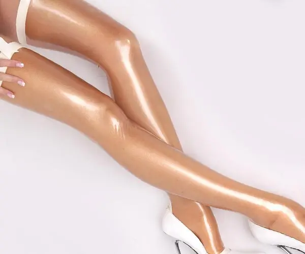 Nahaufnahme von gekreuzten Beinen in glänzenden nackten Strümpfen und weißen High Heels vor einem hellen Hintergrund. Der Glanz der glatten Strümpfe spiegelt die glatte Oberfläche wider, die man nur bei Latex sieht.