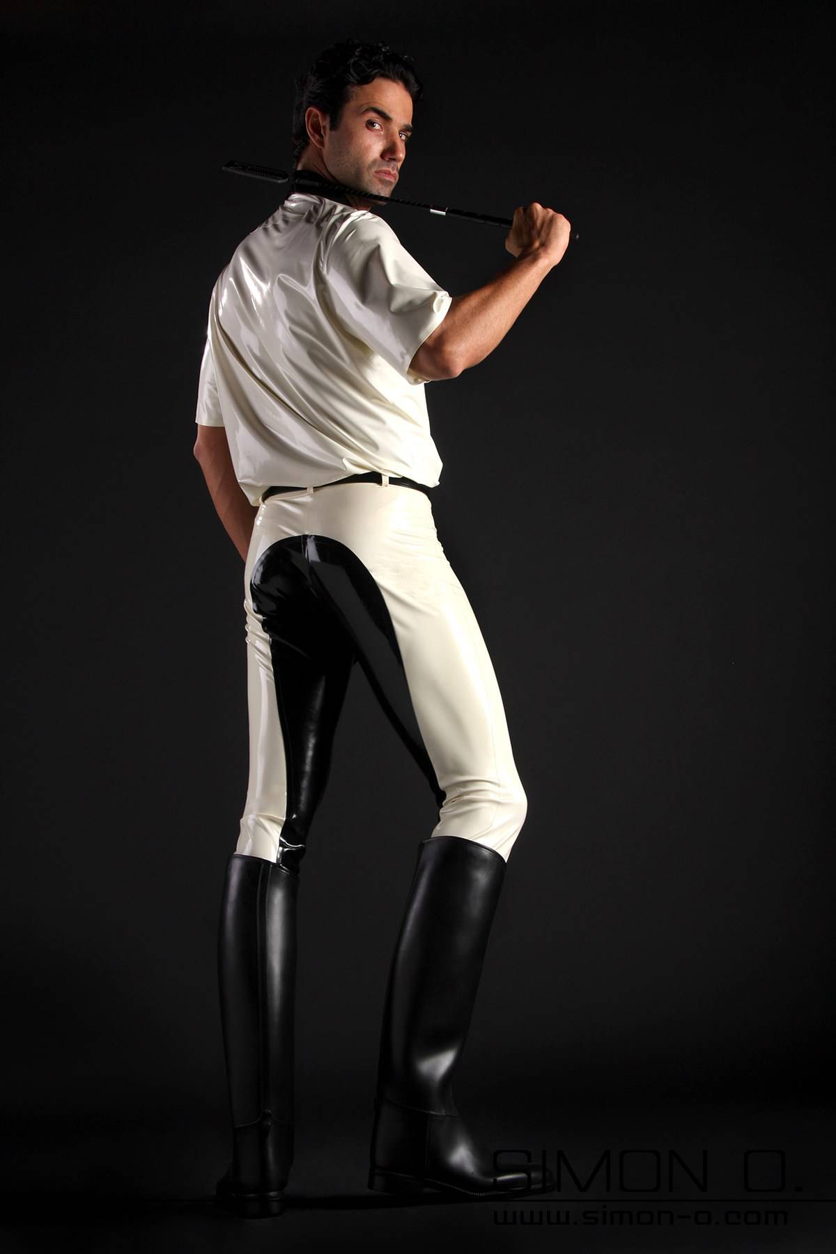 Latex riding breeches men A man with a white latex shirt and a riding crop wears black and white latex breeches and riding boots - seen from behind.