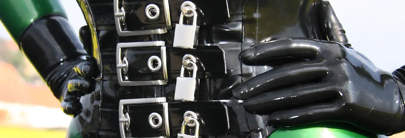 Nahaufnahme einer Person in einem glänzenden Latexkorsett mit drei absperrbaren Vorhängeschlössern in Chrom, eine Hand in Latex Handschuhen ruht auf ihrer Hüfte.