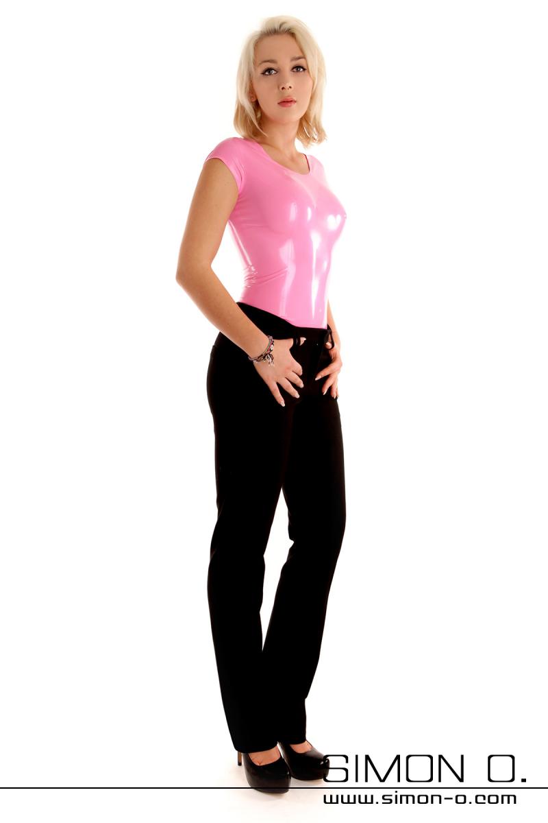 Une femme blonde avec un haut en latex rose à manches courtes et un pantalon noir simple sur un fond blanc.