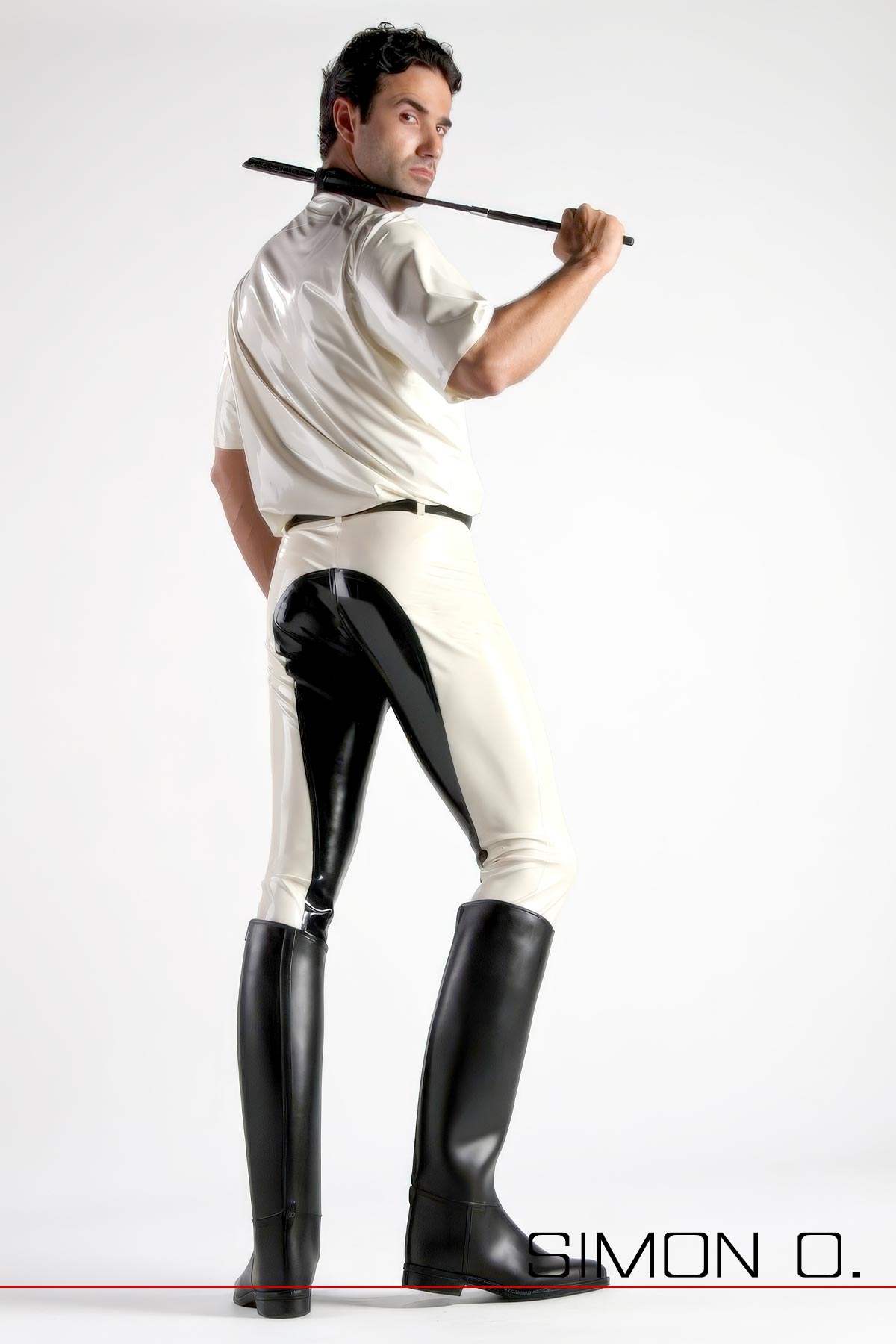 Ein Mann mit weißen Latexhemd und einer Reitgerte trägt eine schwarz weiße Latex Reithose und Reitstiefel - von hinten gesehen.