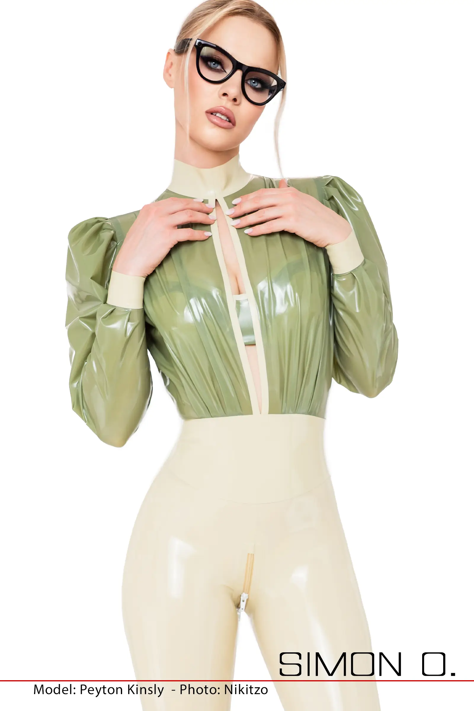 Eine Frau posiert in einem glänzenden, hochtaillierten cremefarbenen Latex Catsuit mit einer hellgrünen Bluse mit Schlitz und Puffärmeln und trägt eine schwarze Katzenaugenbrille vor einem weißen Hintergrund.