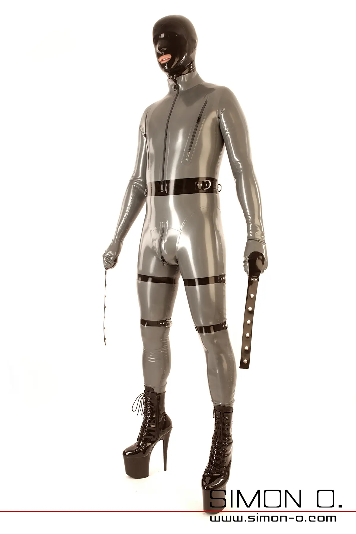 Ein Mann in einem Latex-Bondage-Anzug mit Maske, hochhackigen Stiefeln, Oberschenkelriemen und Fäustlingen mit Fesselriemen von vorne gesehen.