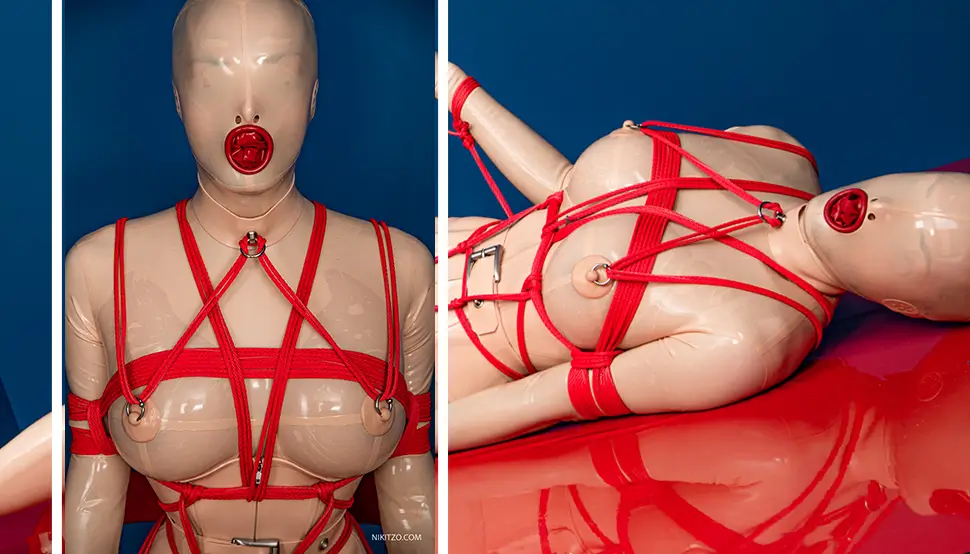 A person in a Latexanzug and a mask is tied up in complicated latex bondage with a red rope and lies on a red surface in front of a striking blue background.
