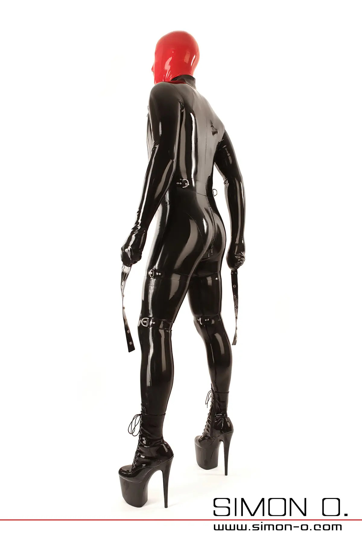 Eine Person in einem schwarzen Bondage Latex-Catsuit mit Fäustlingen und zahlreichen Latex Riemen mit D-Ringen, einer roten Maske, hohen Plateaustiefeln steht vor weißem Hintergrund. 