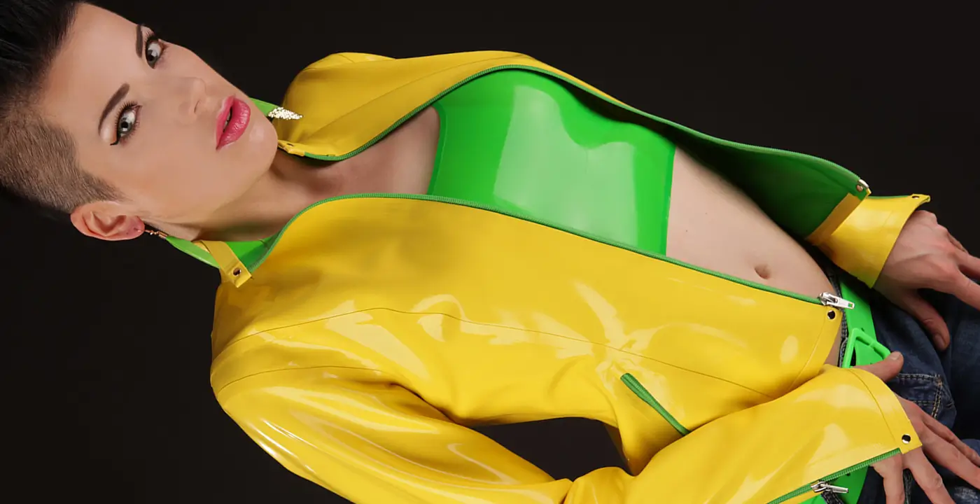 Person, die eine glänzende gelbe Latexjacke über einem glänzenden grünen Oberteil trägt und vor einem dunklen Hintergrund posiert.