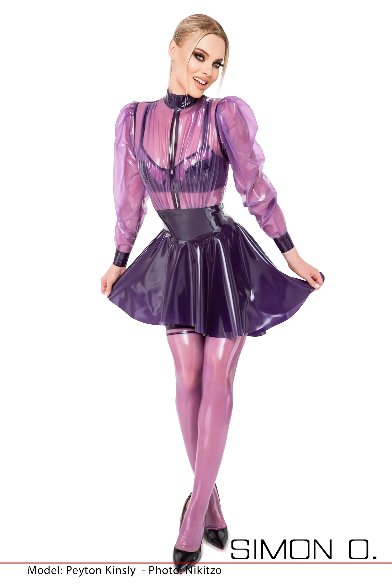 A woman poses in a transparent, purple latex blouse with puffed sleeves and matching stockings in front of a white background.