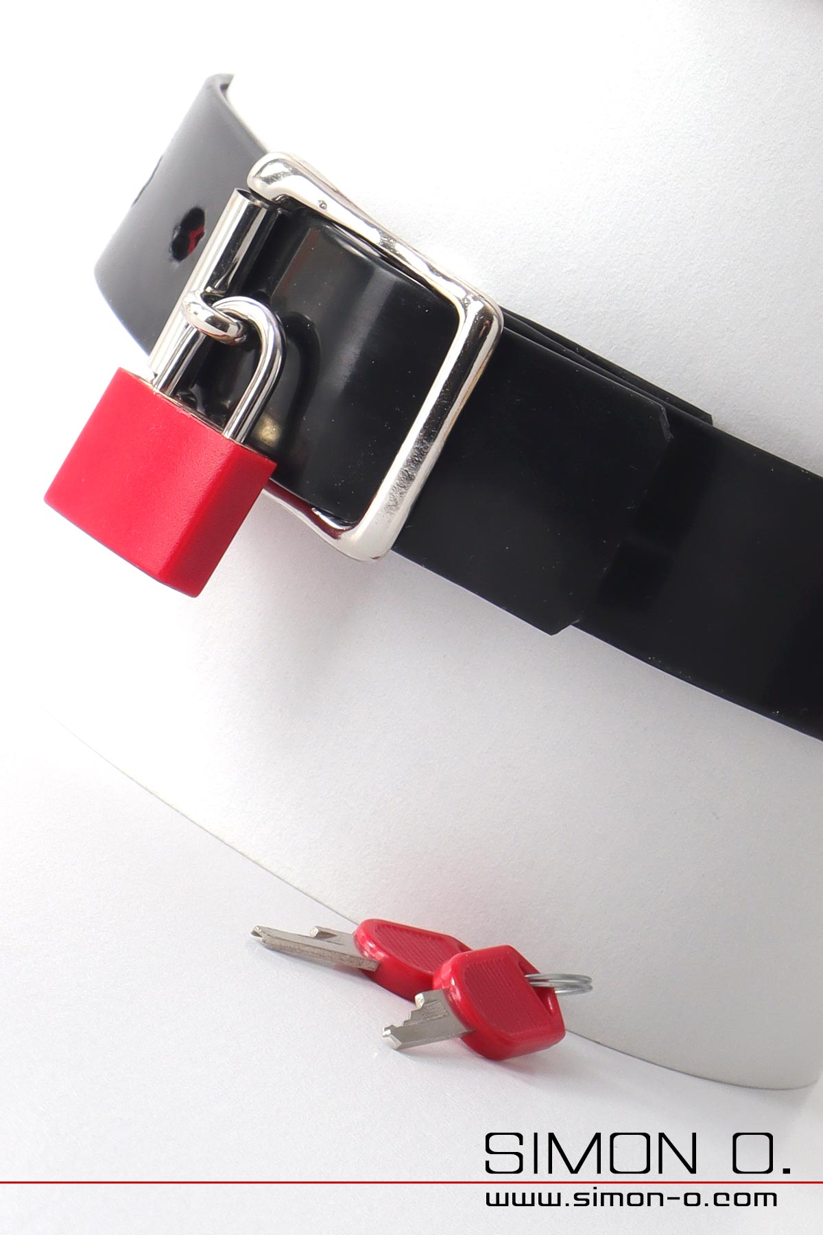 Latex Halsband mit Vorhängeschloss Zu sehen ist ein schwarzes Latexhalsband mit silberner Schnalle und rotem Vorhängeschloss. Auf der weißen Oberfläche darunter liegen zwei Schlüssel.