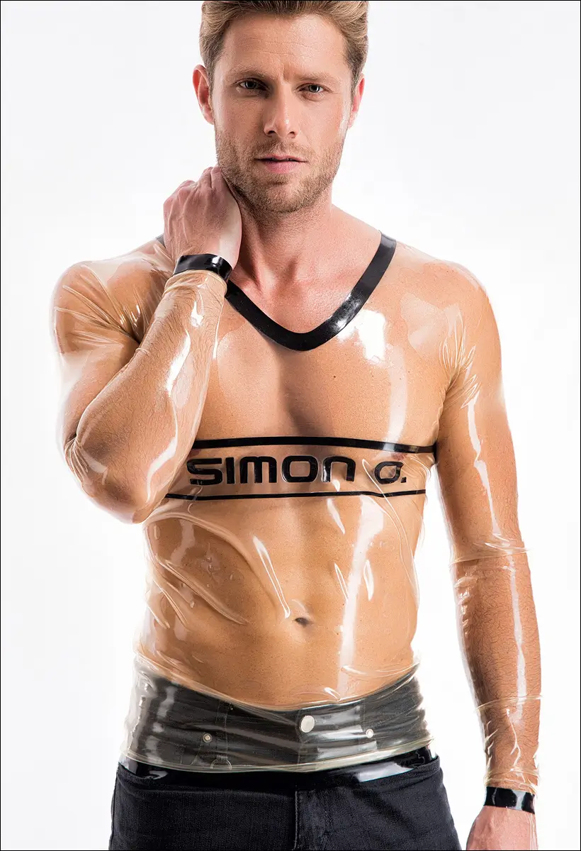 Vor einem weißen Hintergrund steht ein Mann, der ein durchsichtiges, eng anliegendes Herren-Latex-Shirt mit schwarzem Besatz und dem Aufdruck „SIMON O.“ auf der Brust trägt.