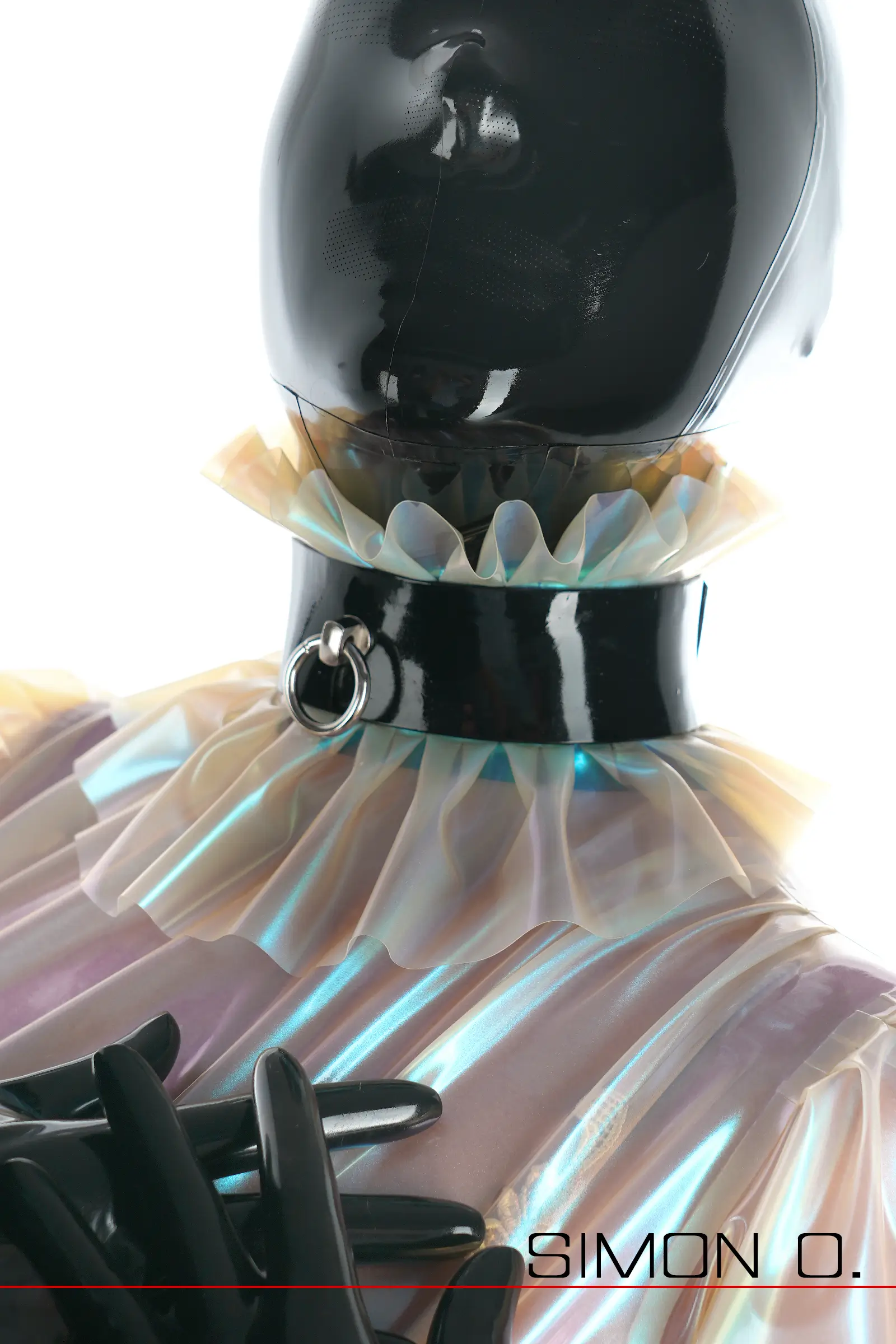 Eine Person mit einer glänzenden schwarzen Maske und einem Latex-Halsband mit O-Ring, die ein durchscheinendes, gerüschtes Kleidungsstück und Handschuhe trägt.