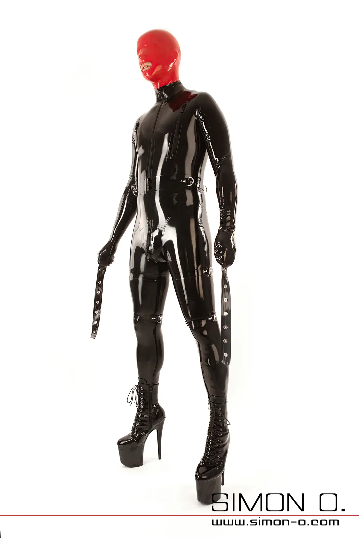 Eine Mann in einem schwarzen Bondage Latex-Catsuit mit Fäustlingen und zahlreichen Latex Riemen mit D-Ringen, einer roten Maske, hohen Plateaustiefeln steht vor weißem Hintergrund von vorne gesehen. 