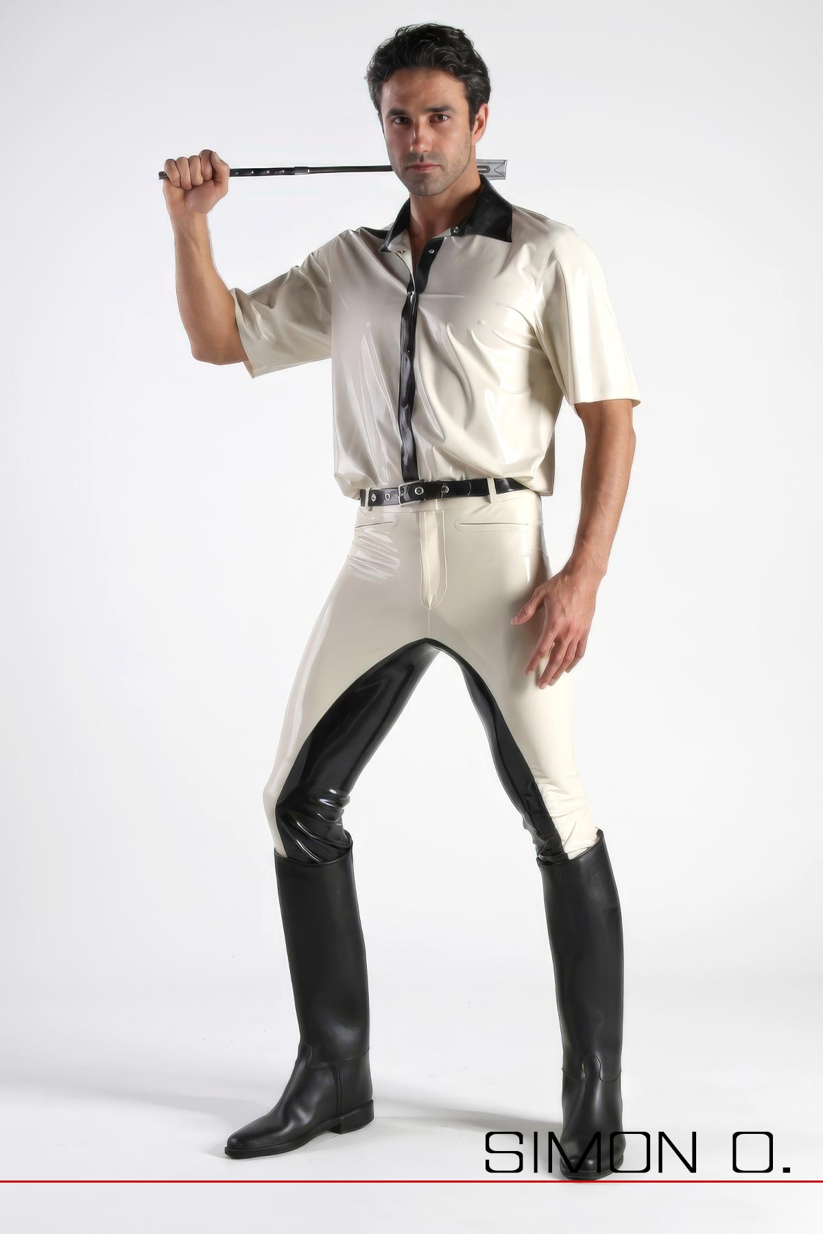 Ein mann mit weißen Latexhemd und einer Reitgerte hinter den Kopf trägt eine schwarz weiße Latex Reithose und Reitstiefel.