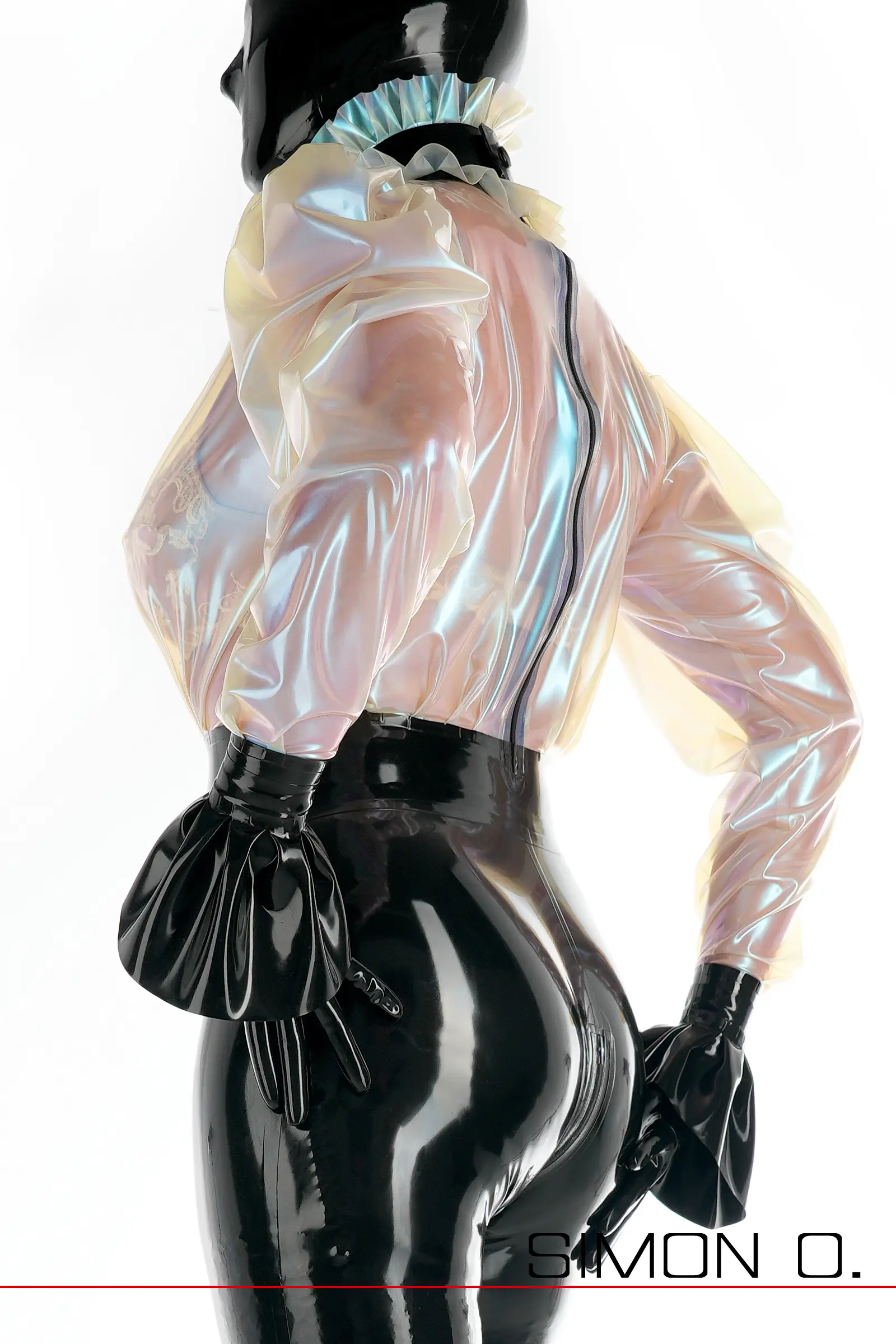 Eine Person, die eine glänzende, schillernde Bluse sowie schwarze Latexhandschuhe und -hosen trägt, von der Seite vor einem weißen Hintergrund gesehen.